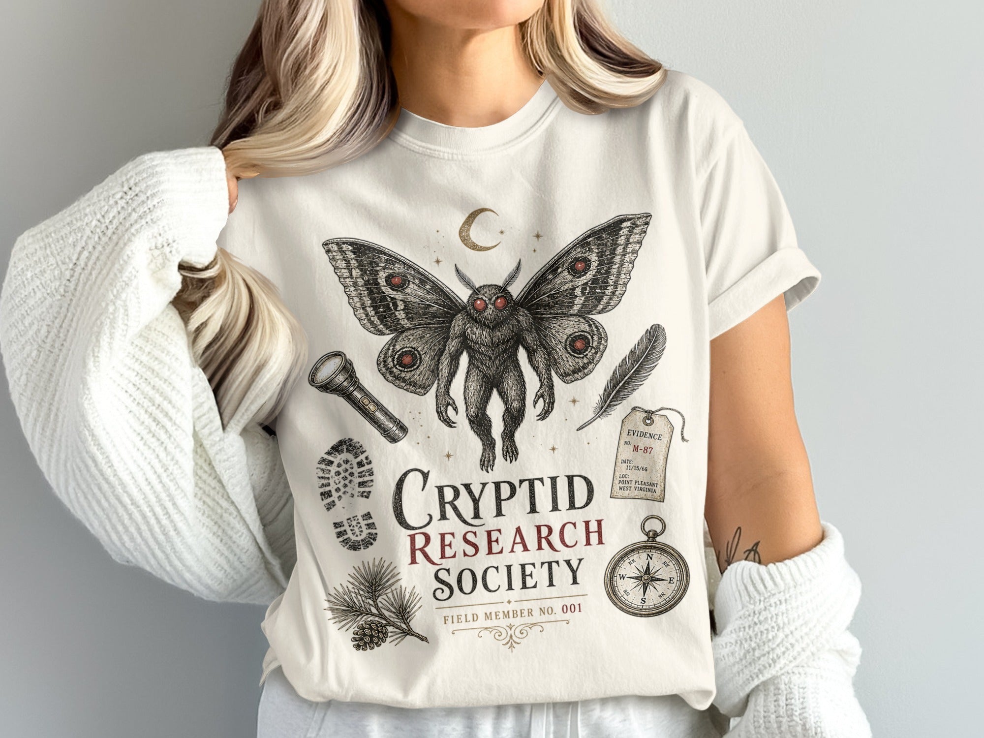Cream t-shirt with mothman, flashlight, footprint, feather, compass, and "Cryptid Research Society" text.