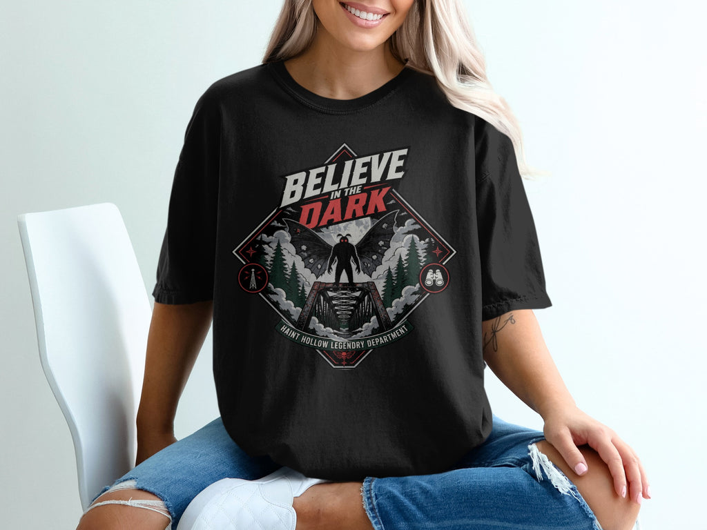 Black t-shirt with "BELIEVE IN THE DARK" and mothman on a bridge graphic.