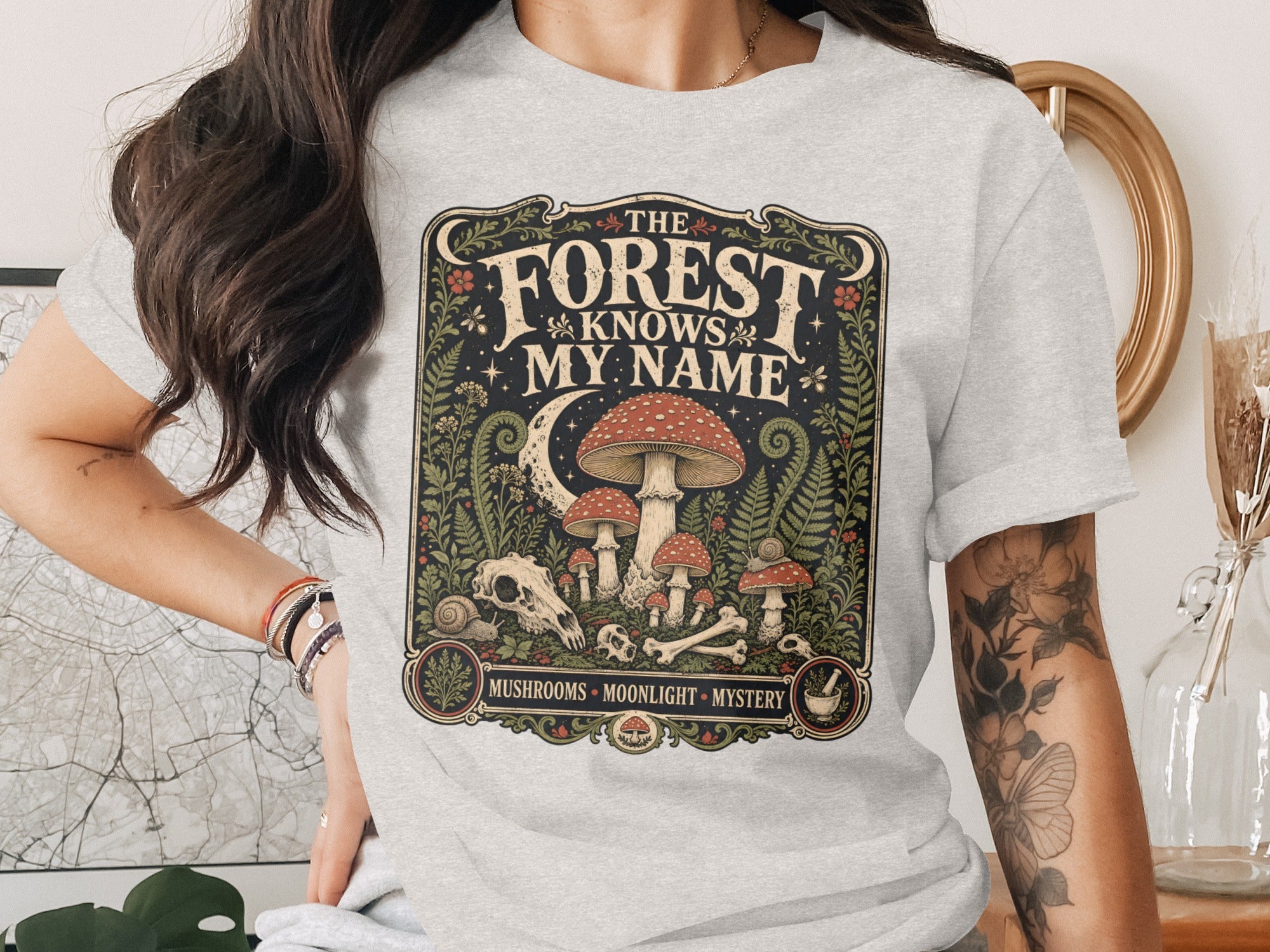 T-shirt graphic with mushrooms, skull, bones, and text "The Forest Knows My Name".