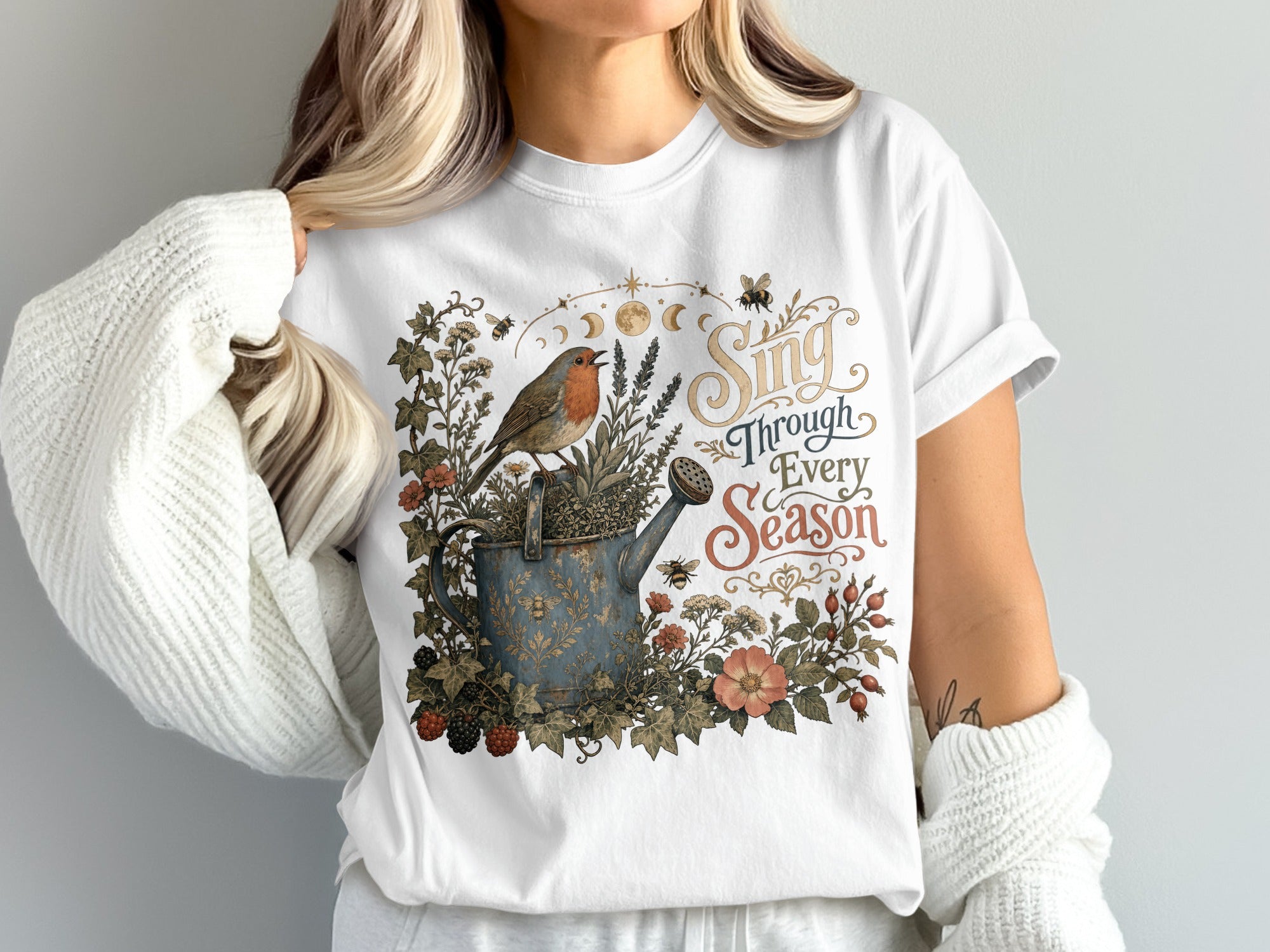 White t-shirt with a robin on a watering can surrounded by foliage and text "Sing Through Every Season."
