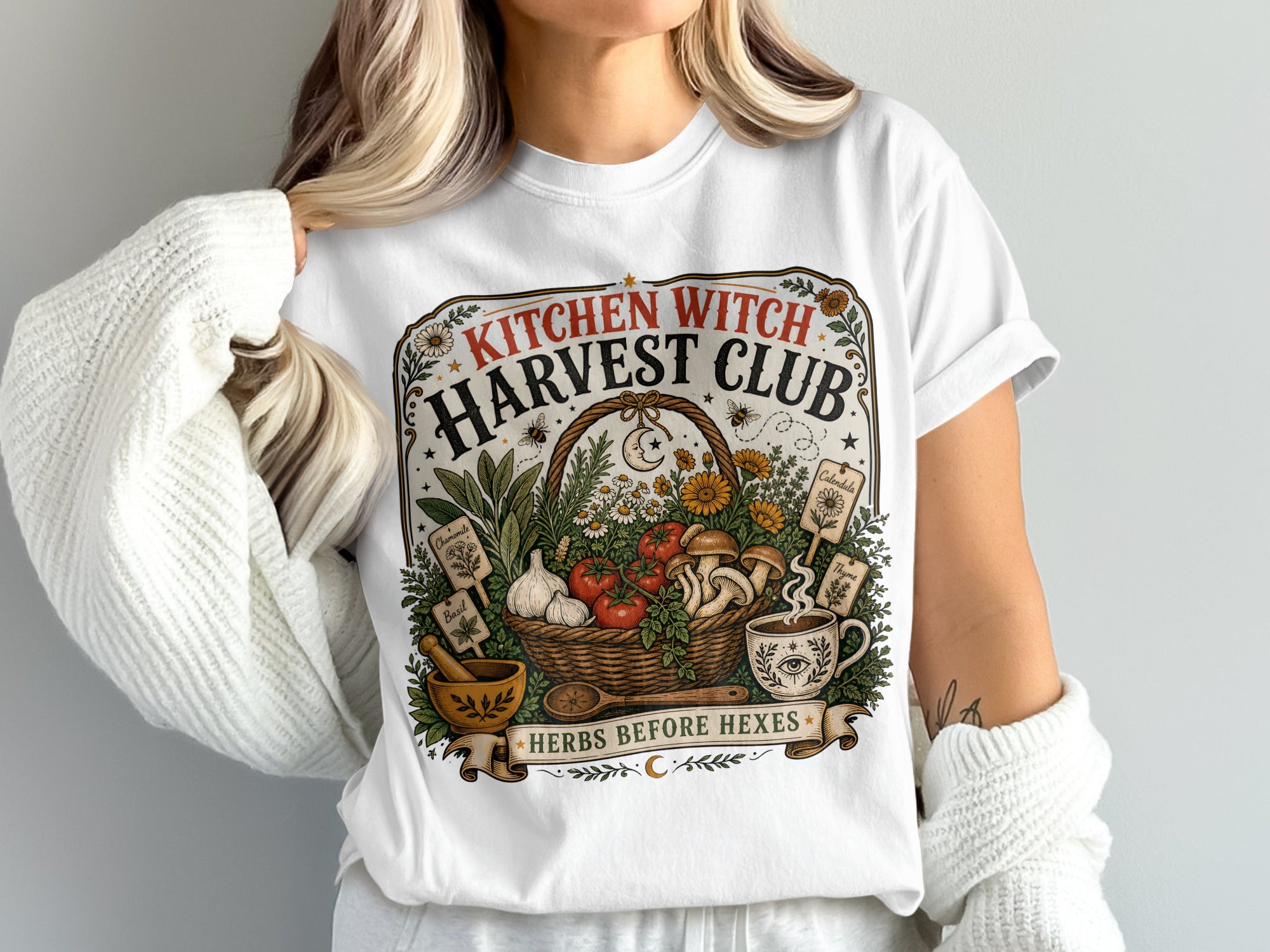 White t-shirt with "Kitchen Witch Harvest Club" graphic featuring harvest basket, herbs, and witchy symbols.
