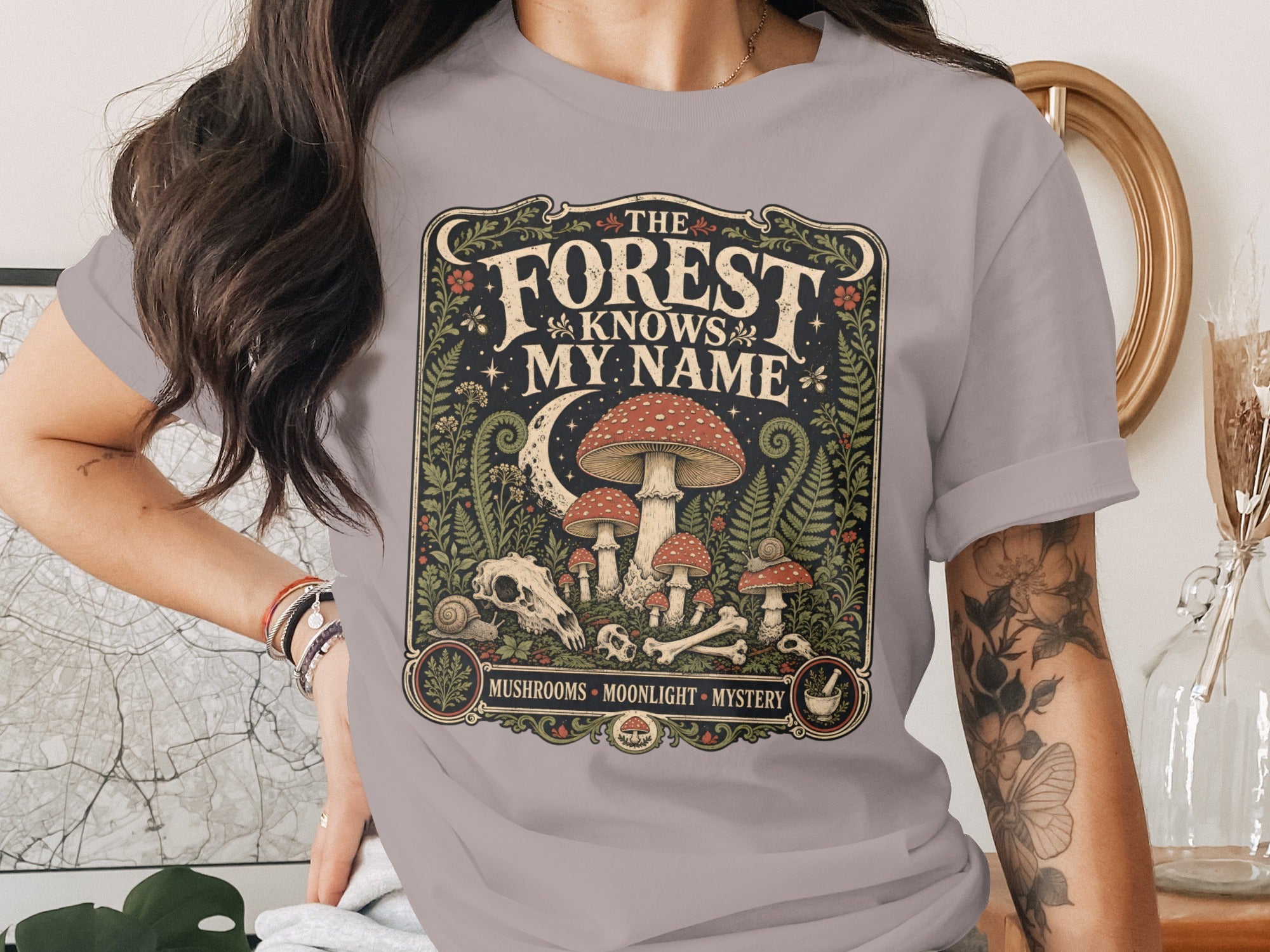 T-shirt graphic with text "The Forest Knows My Name" and mushroom, moon, and skull imagery.