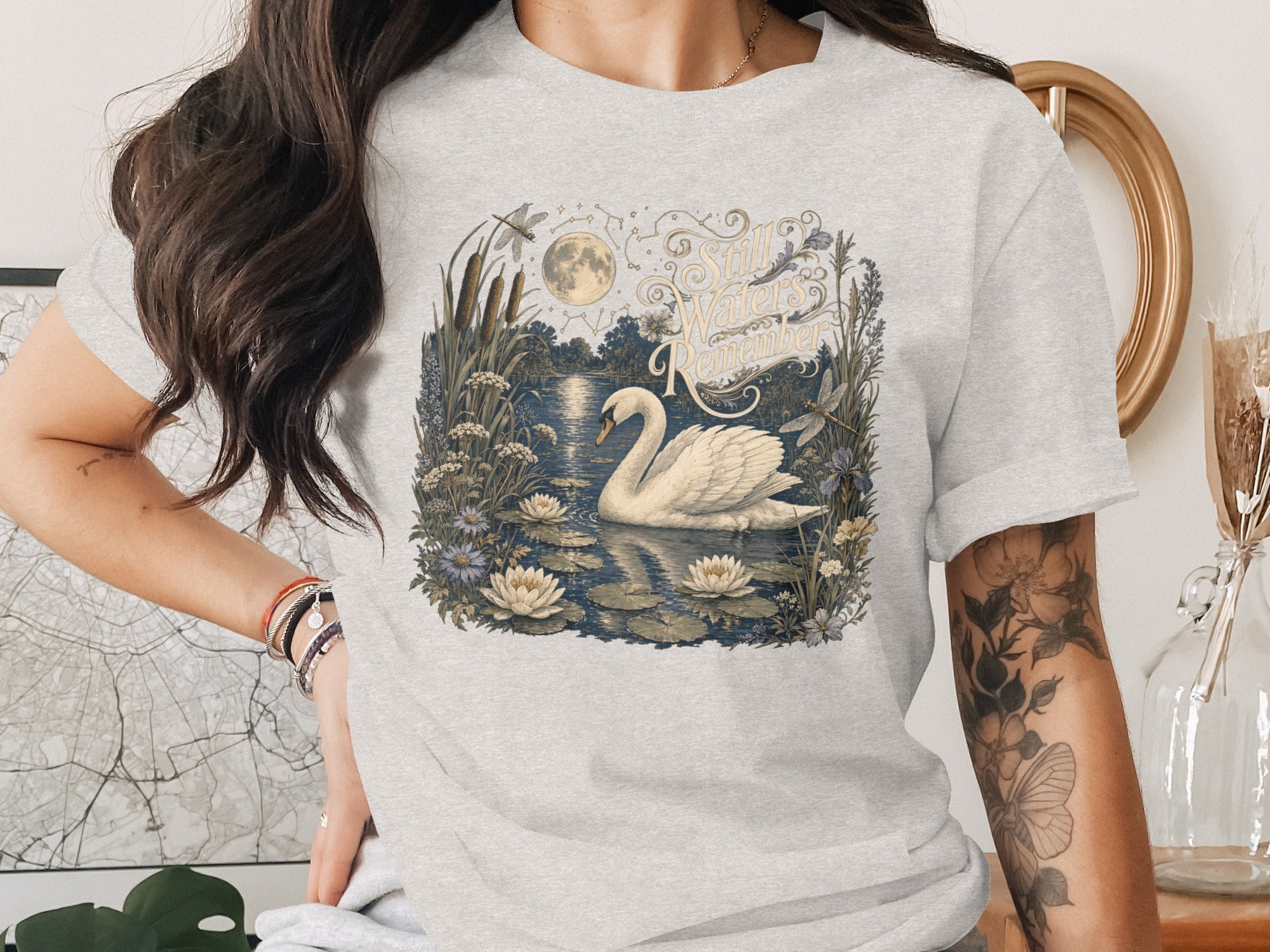 White swan on water with lilies, moon, dragonflies, and text "Still Waters Remember".
