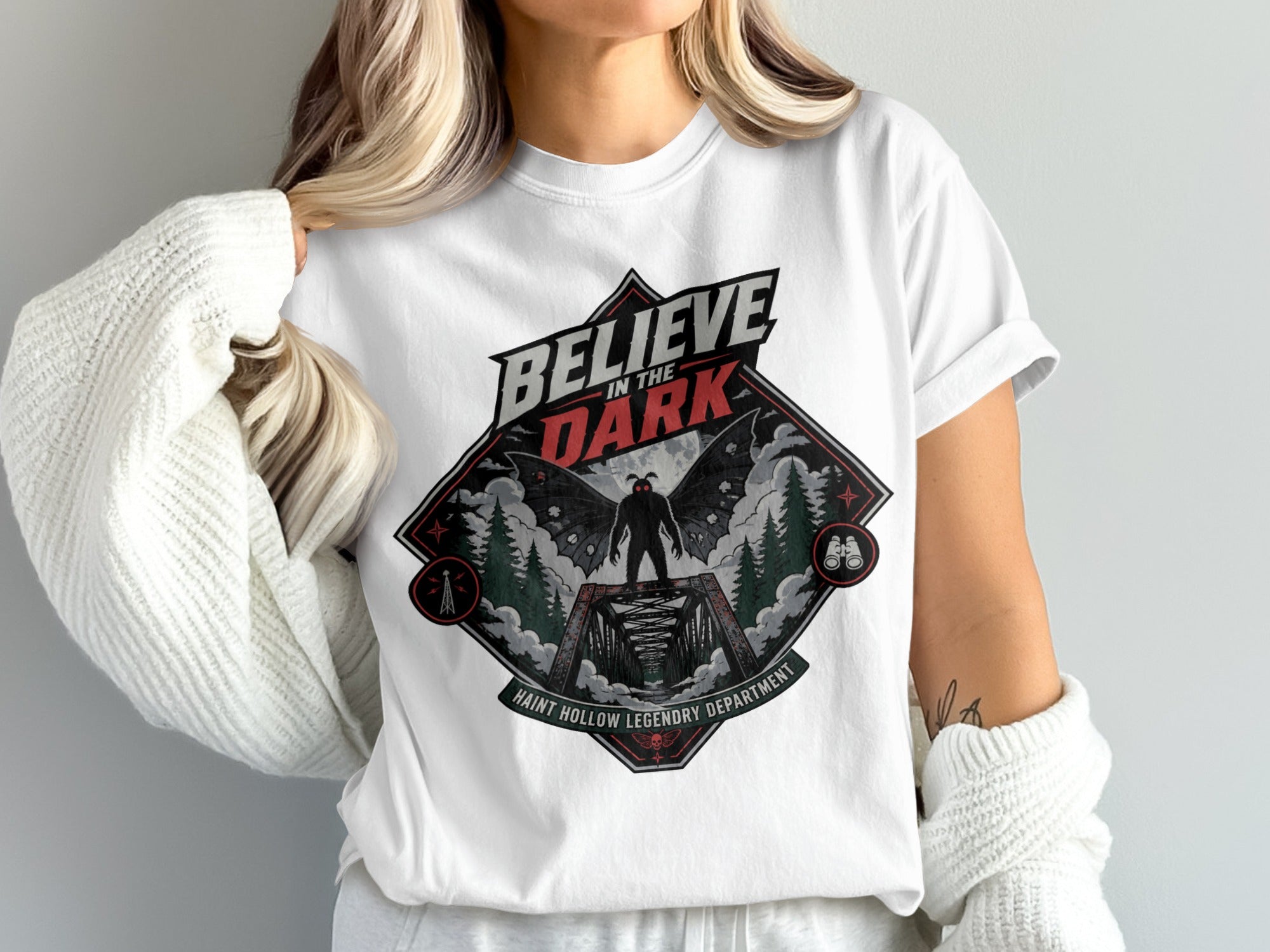 T-shirt graphic with text "Believe in the Dark" and a winged creature on a bridge.