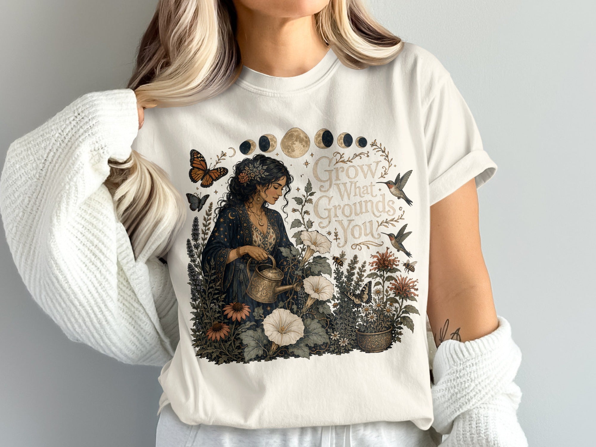 T-shirt with a woman watering plants, celestial moon phases, and the text "Grow What Grounds You."