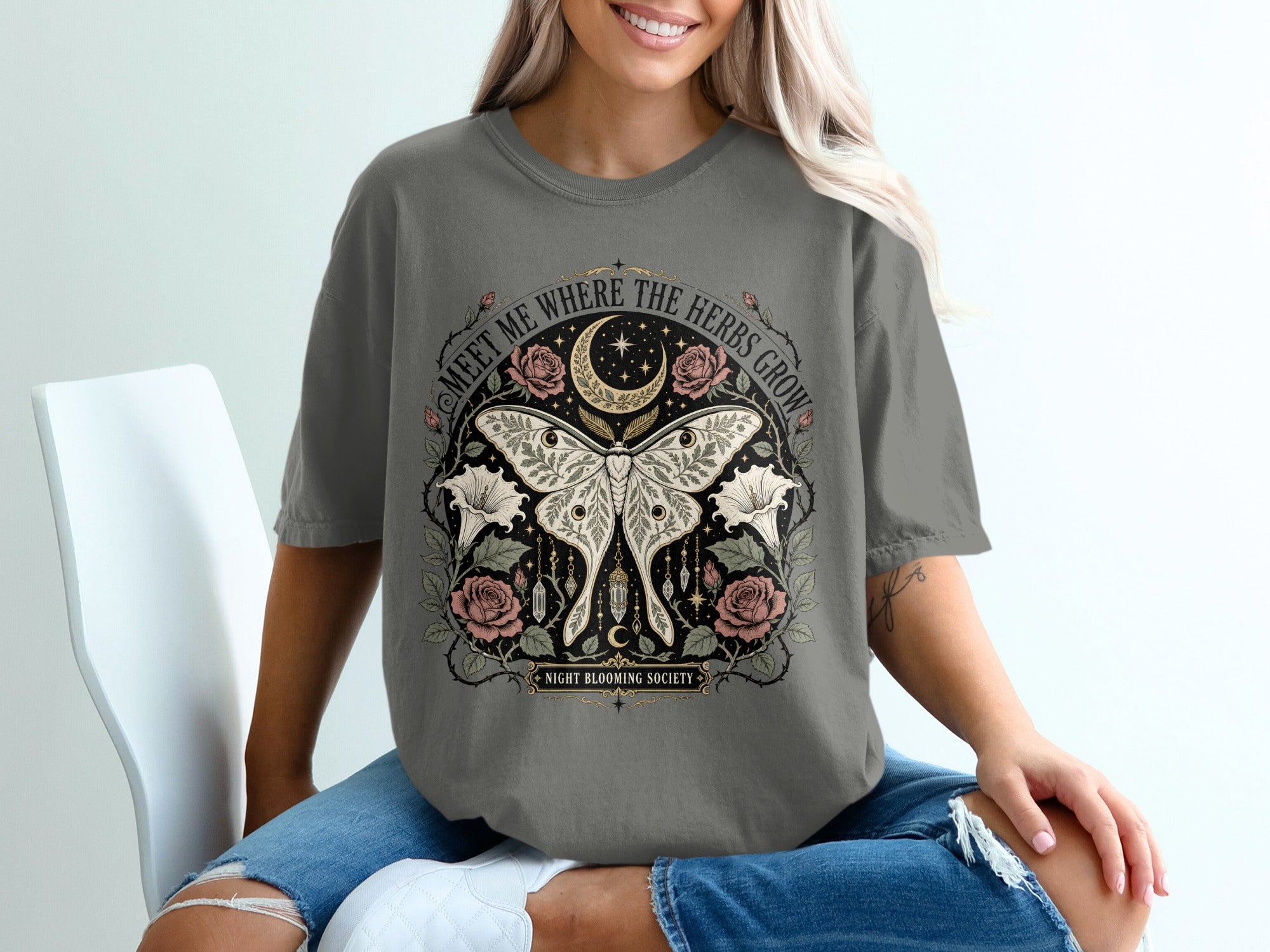 Grey t-shirt with a circular design featuring moths, roses, moon, stars, and the text "Meet me where the herbs grow".