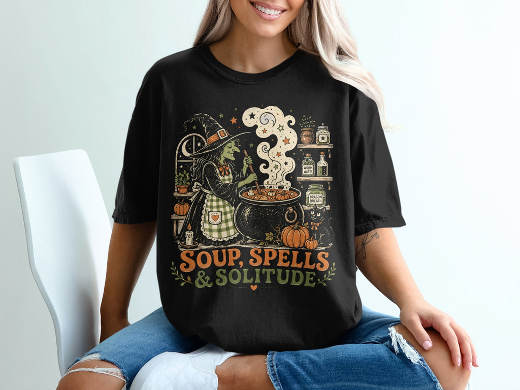 Black t-shirt graphic with a witch stirring a cauldron under the moon, text reads "SOUP, SPELLS & SOLITUDE".