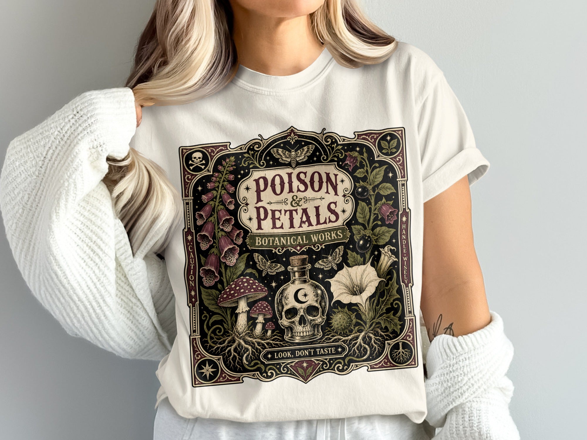 White t-shirt with "Poison & Petals Botanical Works" design featuring skull, bottle, mushrooms, and flowers.