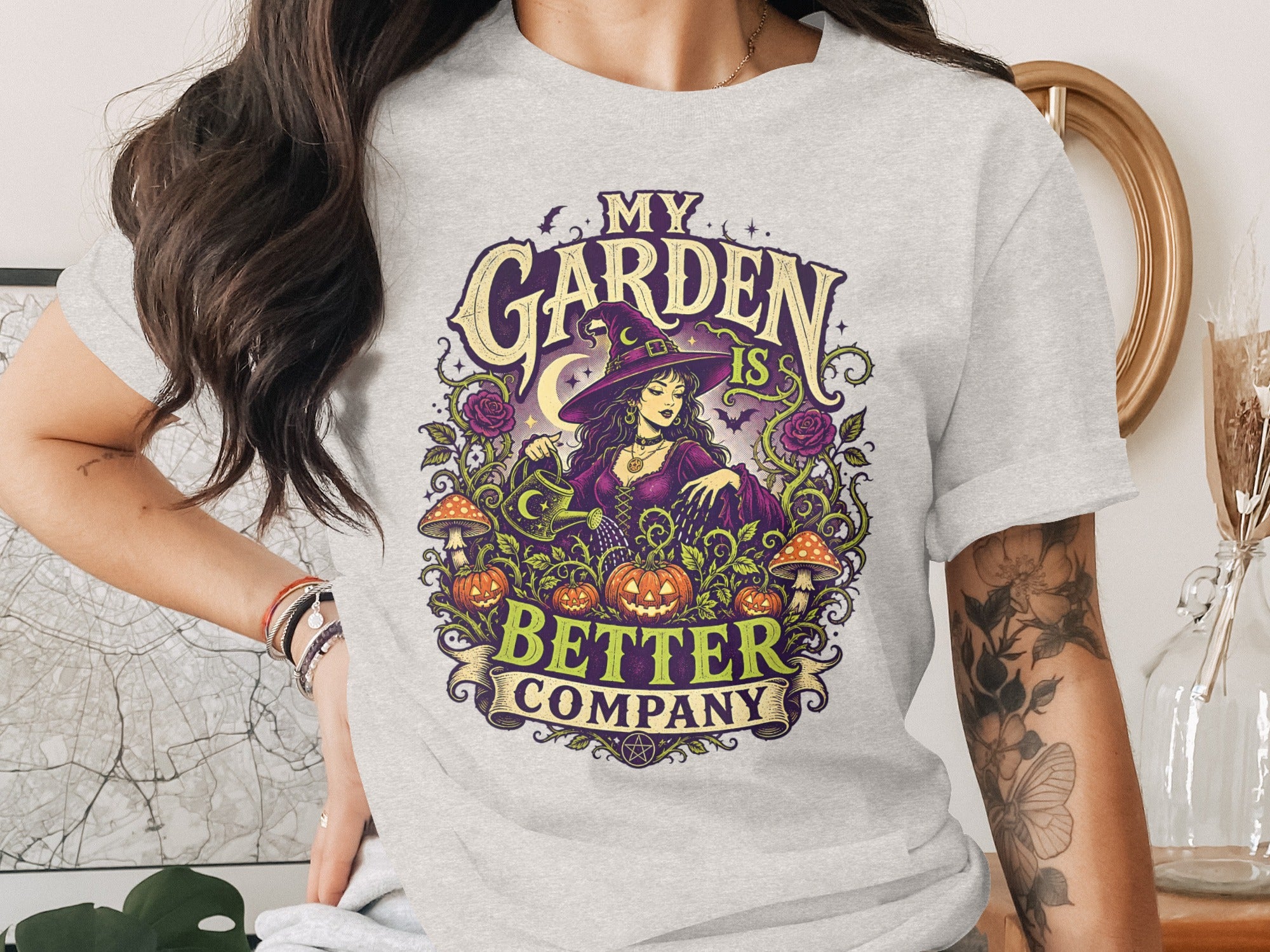 Graphic tee with witch and pumpkins surrounded by plants, reading "My Garden Is Better Company."
