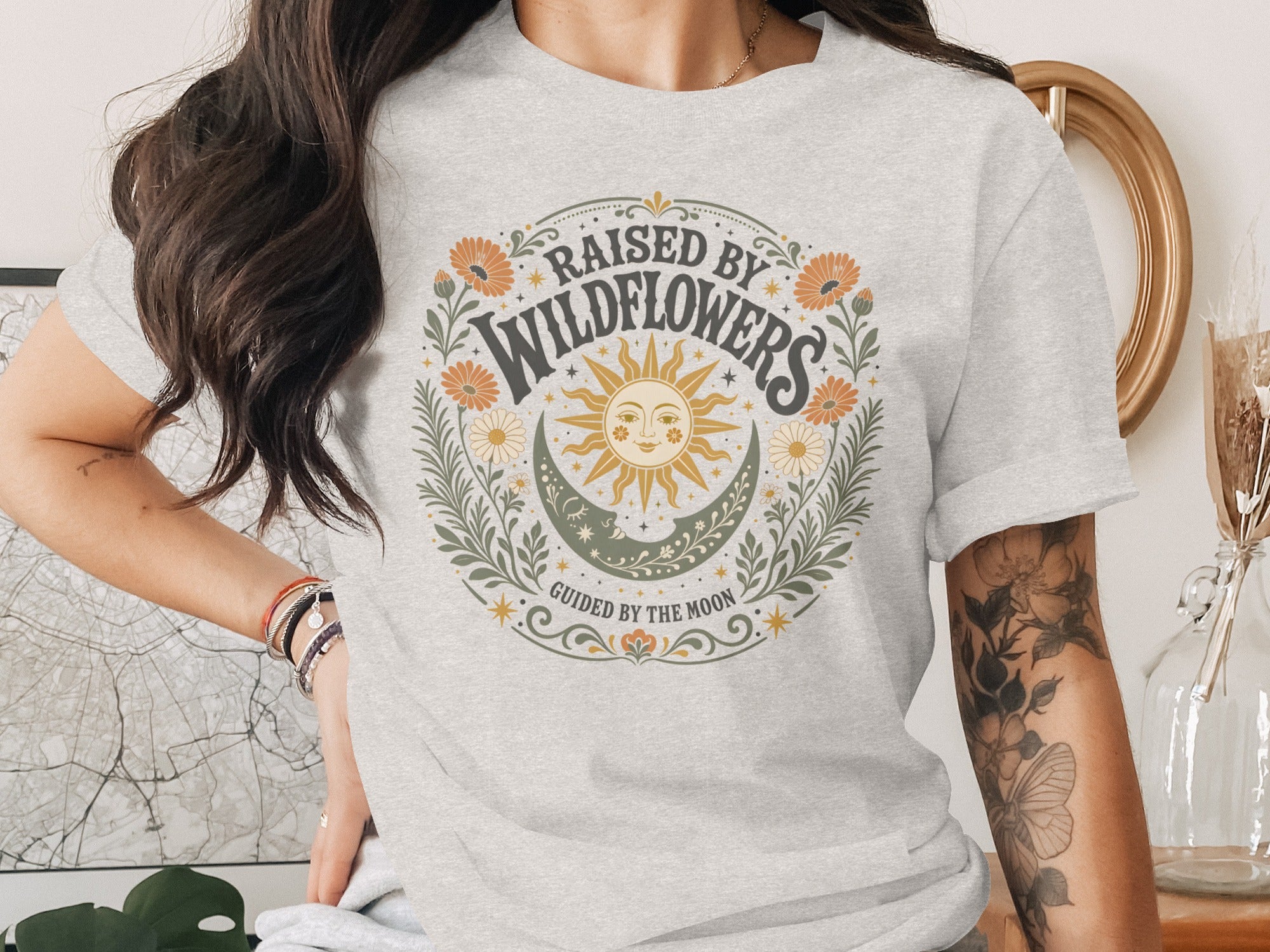 Grey t-shirt with circular graphic: "Raised by Wildflowers" above sun, moon, stars and flowers.
