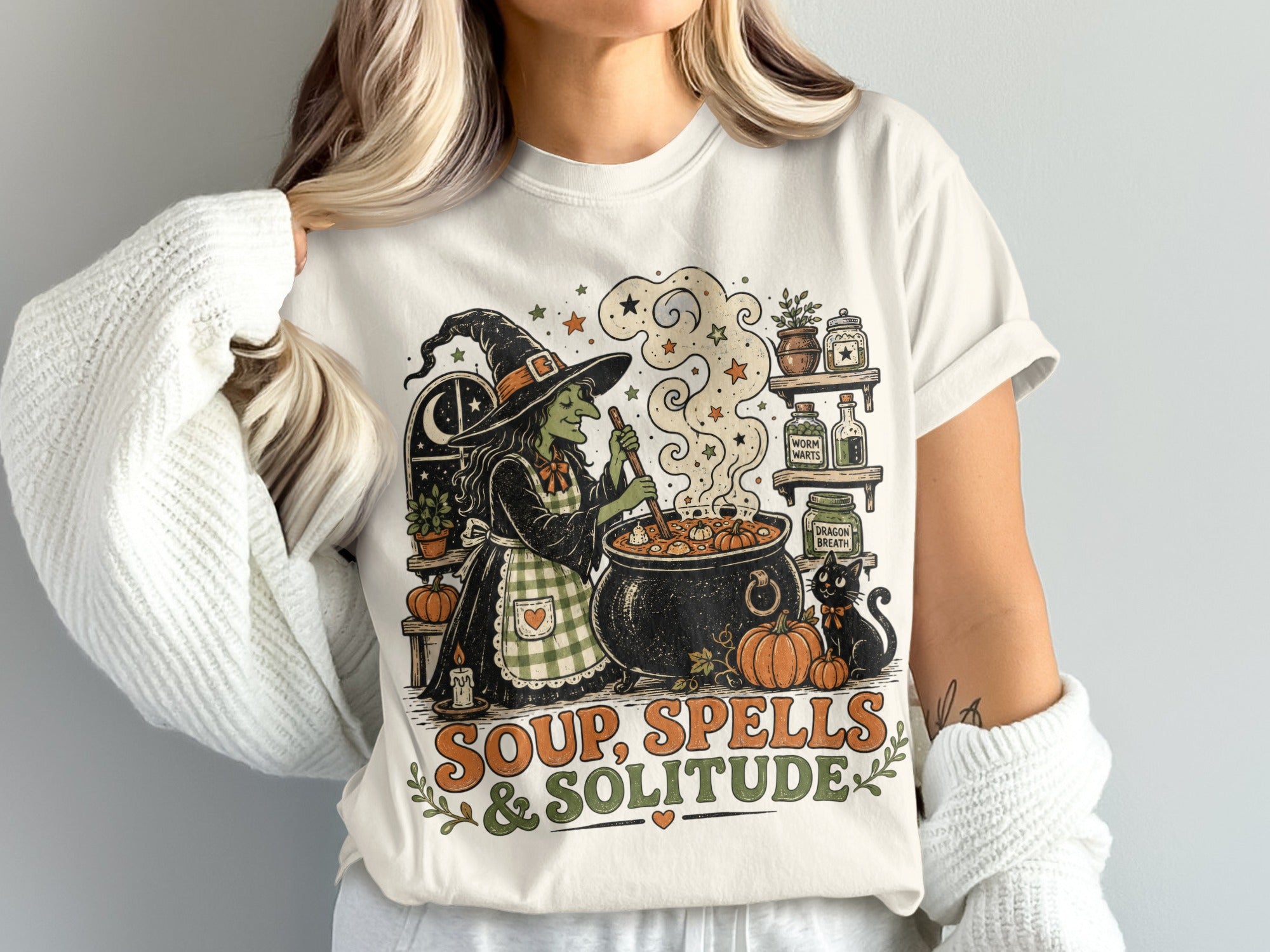 Graphic t-shirt with a witch making soup, a black cat, pumpkins, and text "Soup, Spells & Solitude".
