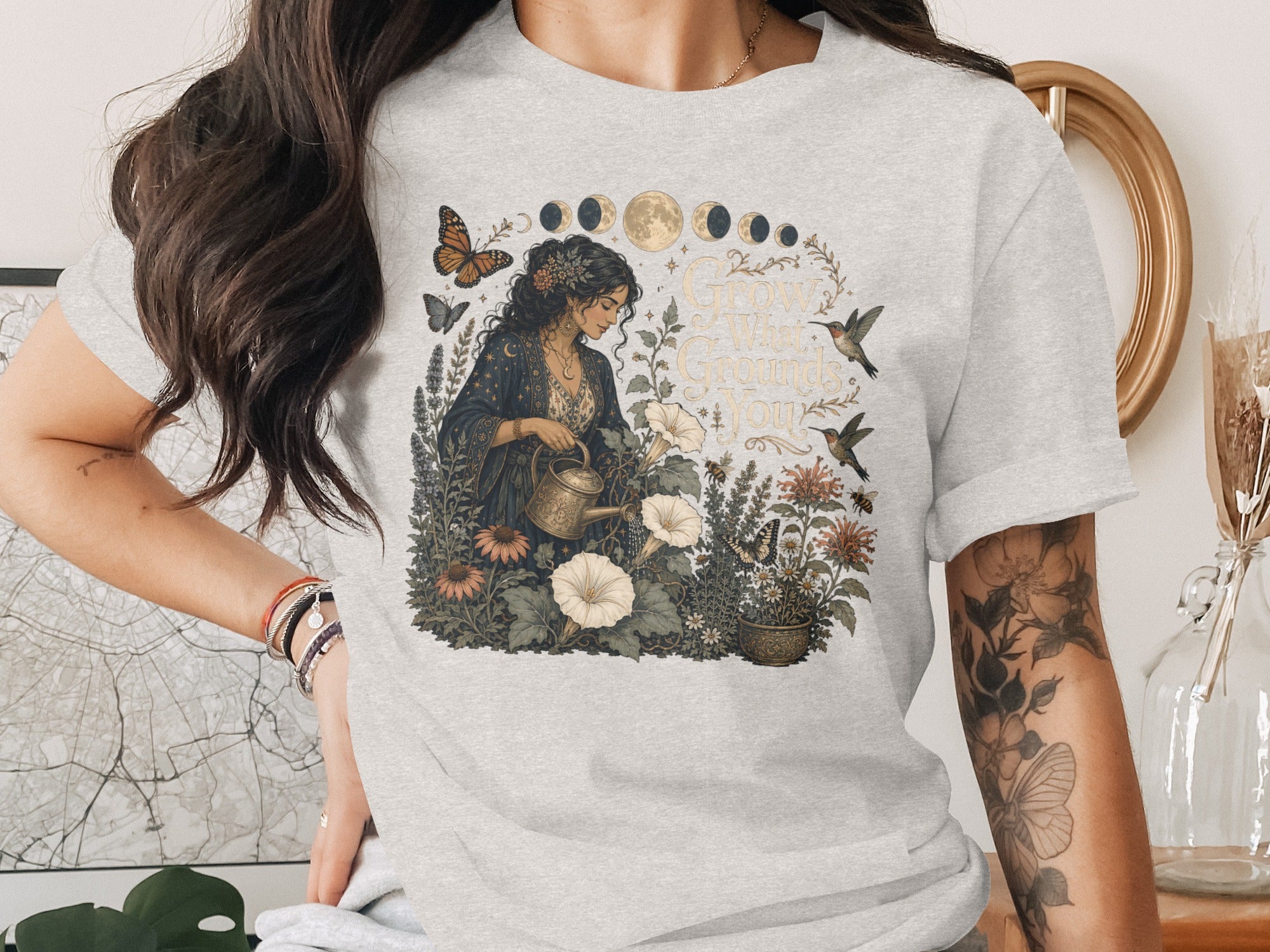 T-shirt graphic with woman watering plants, butterflies, hummingbirds, moon phases, and text.