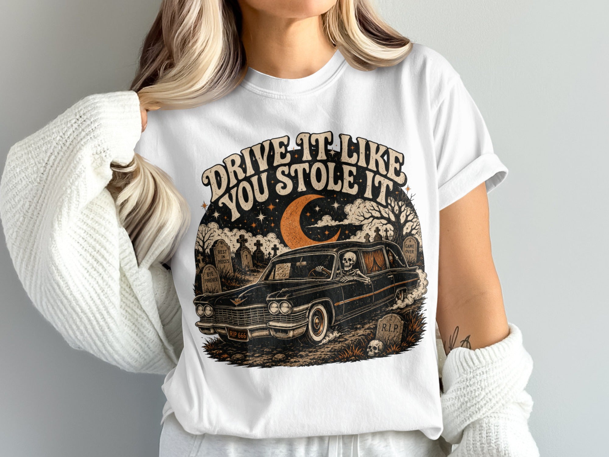T-shirt graphic: Skeleton driving hearse through cemetery, "Drive It Like You Stole It" text.