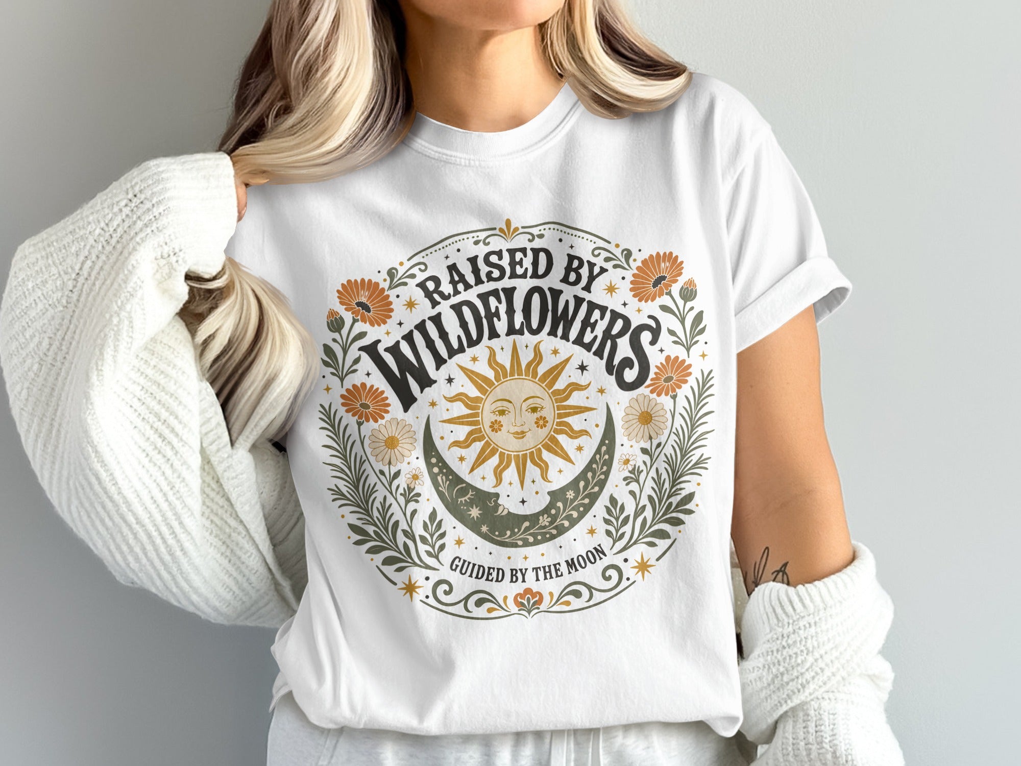 White t-shirt with a circular floral design featuring "Raised by Wildflowers" and a sun and moon graphic.