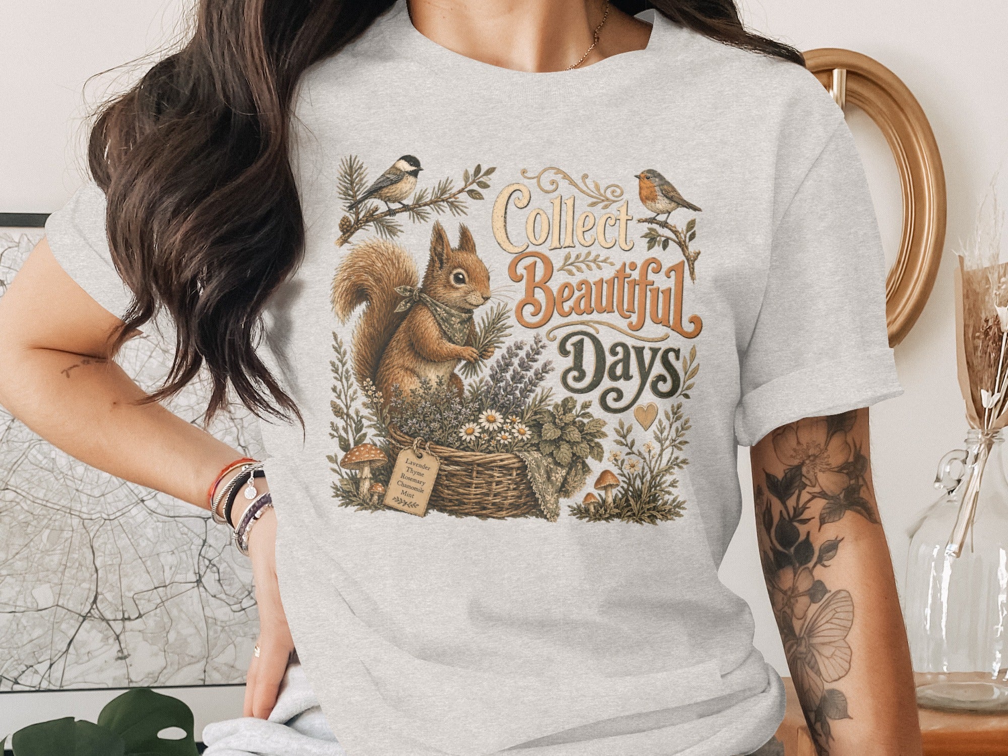 Light grey t-shirt with squirrel, birds, and basket of herbs graphic with text "Collect Beautiful Days".