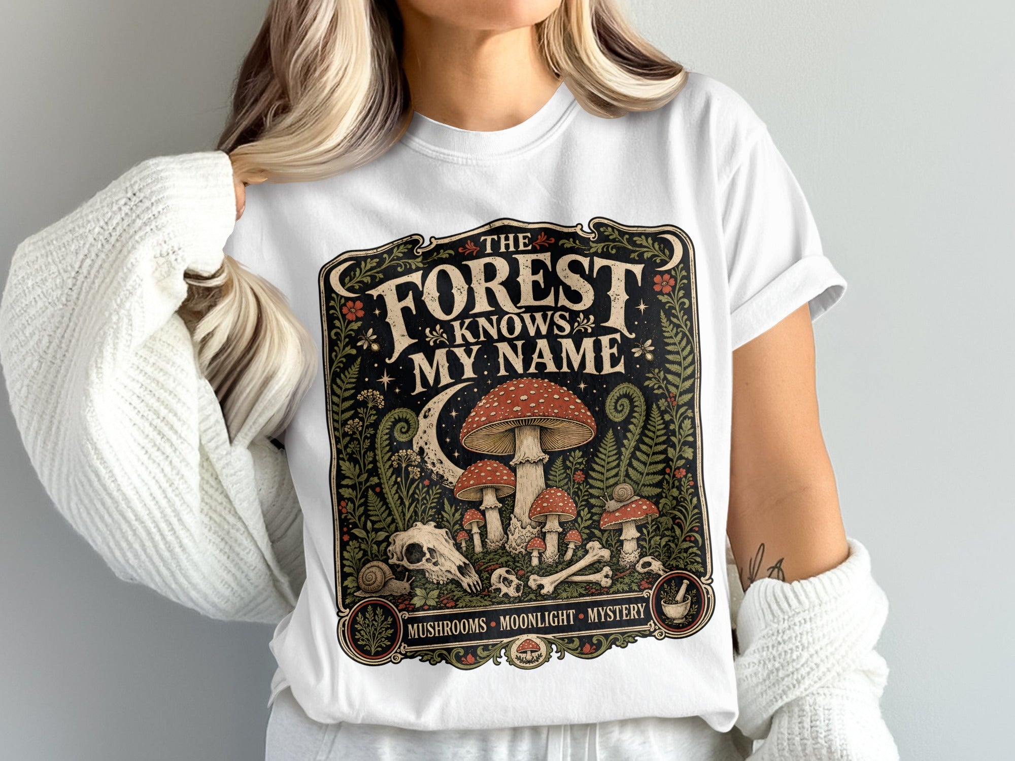 White t-shirt with "The Forest Knows My Name" graphic featuring mushrooms, moon, and bones.