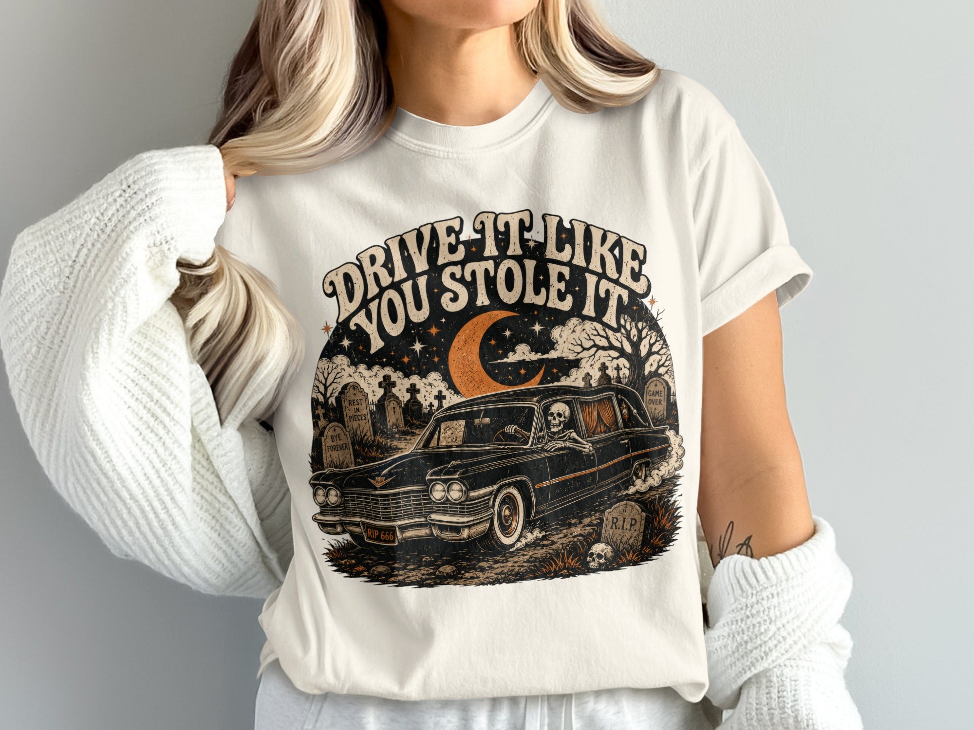 T-shirt graphic with skeleton driving hearse through cemetery, "Drive It Like You Stole It".