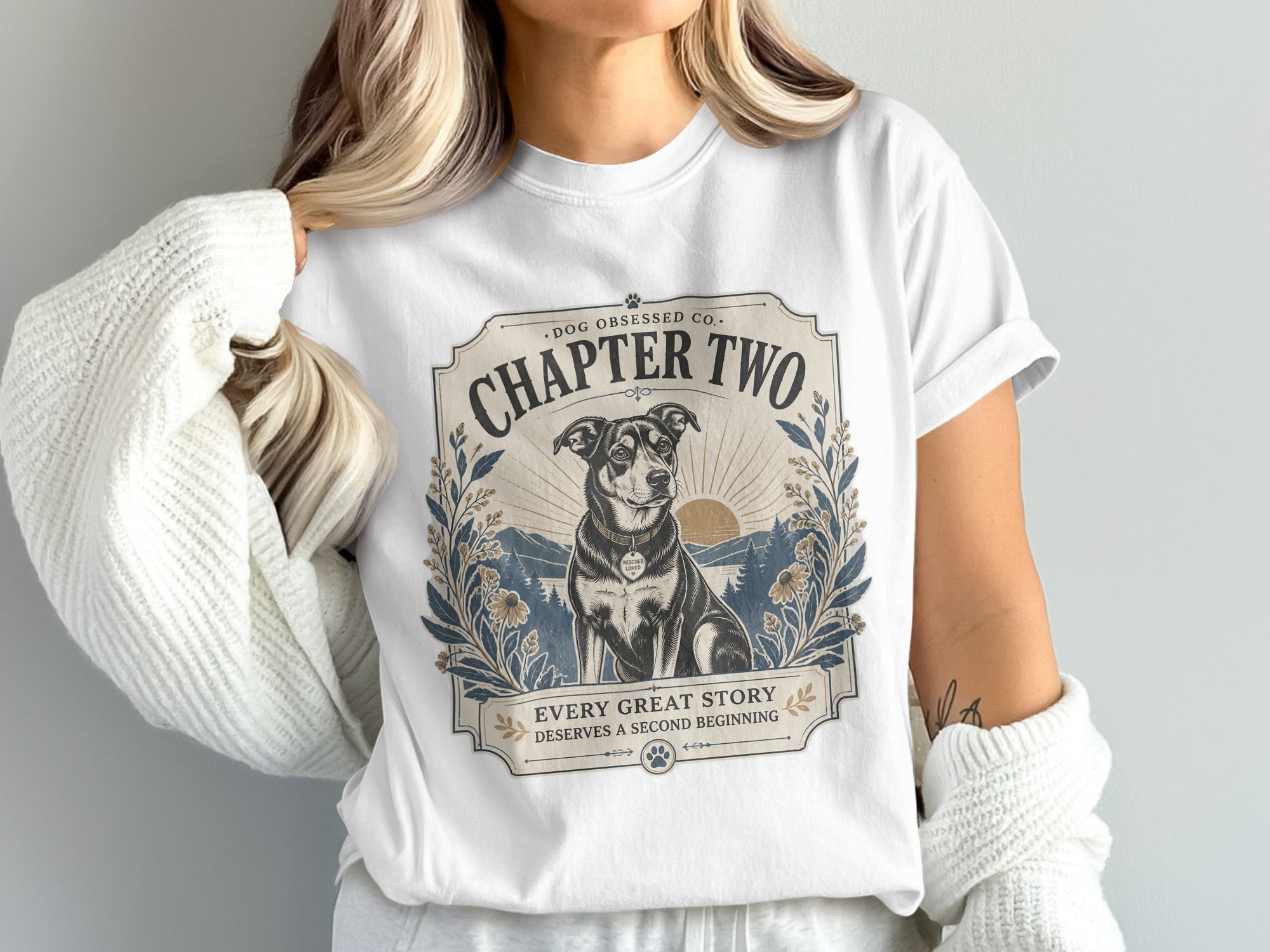 White t-shirt featuring a framed design with a dog, mountains, and the text "Chapter Two" and "Every Great Story Deserves A Second Beginning."