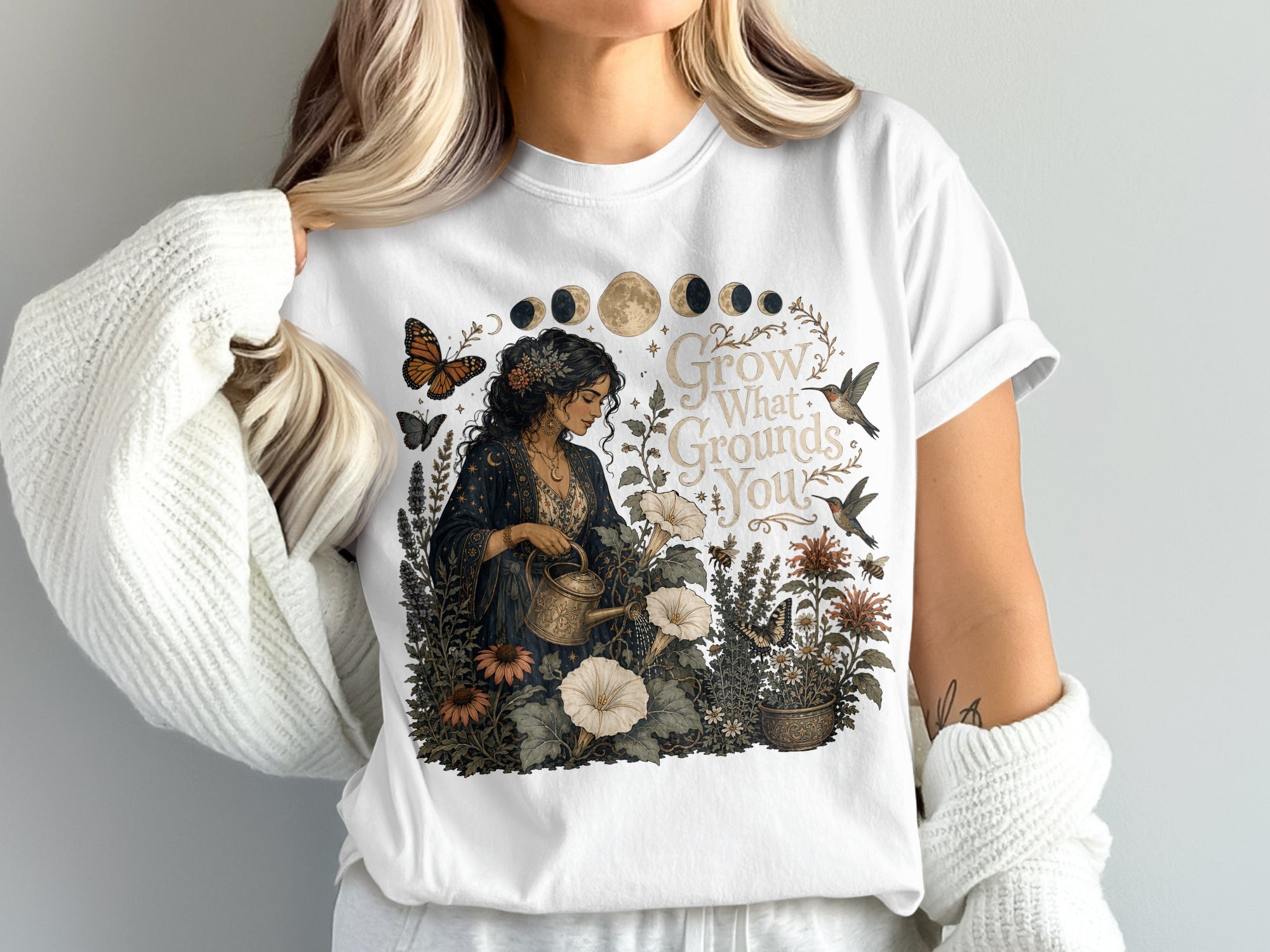 Graphic tee with woman watering plants, text "Grow What Grounds You", moon phases, butterflies, and hummingbirds.