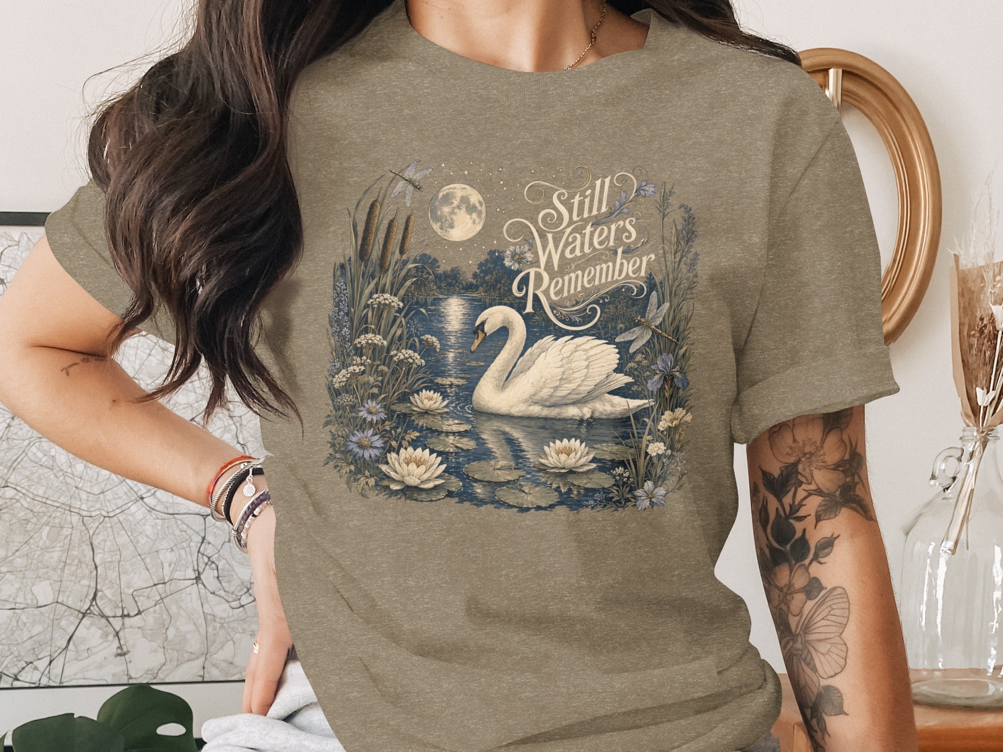 T-shirt graphic with swan, water lilies, moon, dragonflies, and text "Still Waters Remember".