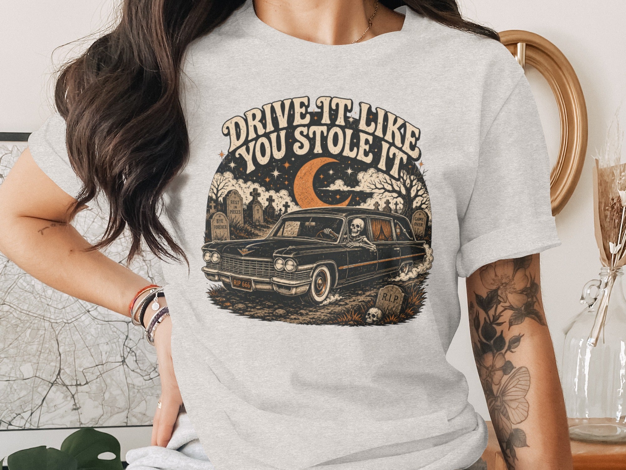 T-shirt graphic with vintage car, skeleton driver, cemetery scene, and text "DRIVE IT LIKE YOU STOLE IT".