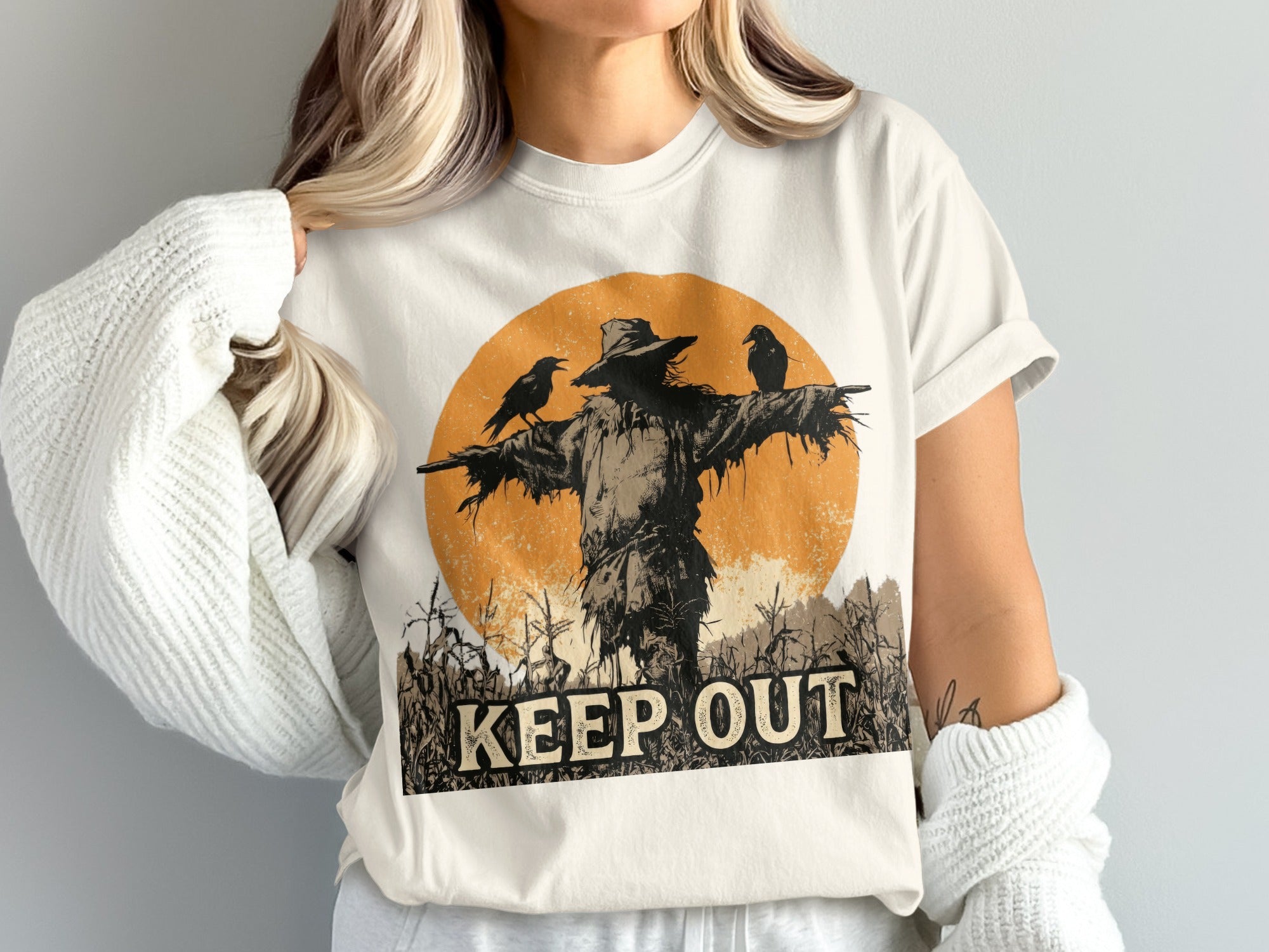 Scarecrow and crows graphic with "KEEP OUT" text on a light-colored t-shirt.