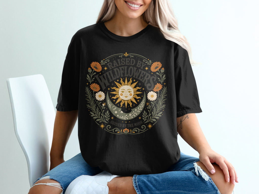 Black t-shirt graphic with sun, moon, wildflowers, and text "RAISED BY WILDFLOWERS" and "GUIDED BY THE MOON".