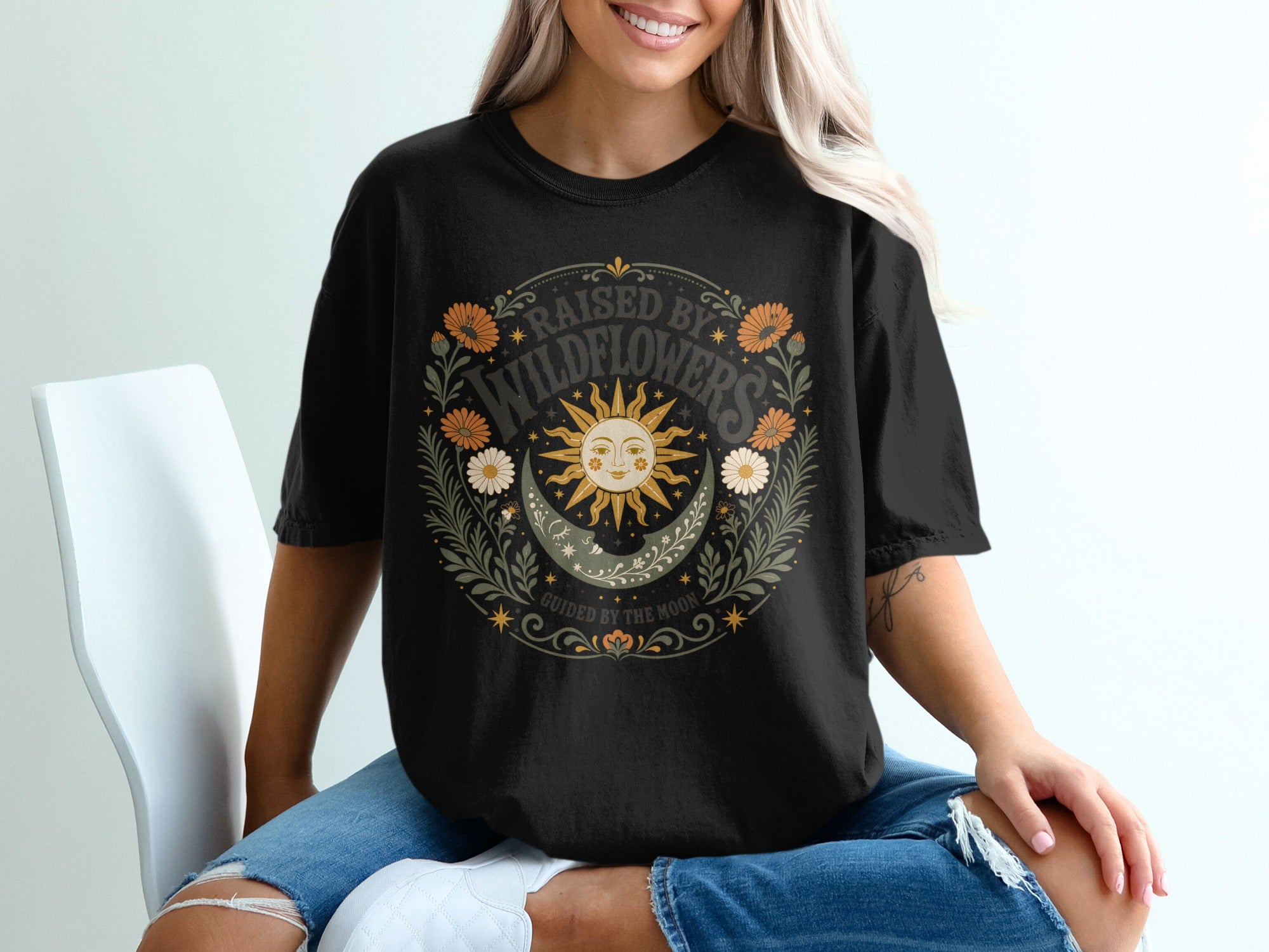 Black t-shirt graphic with sun, moon, wildflowers, and text "RAISED BY WILDFLOWERS" and "GUIDED BY THE MOON".