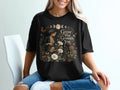 Black t-shirt with woman watering plants and "Grow What Grounds You" text.