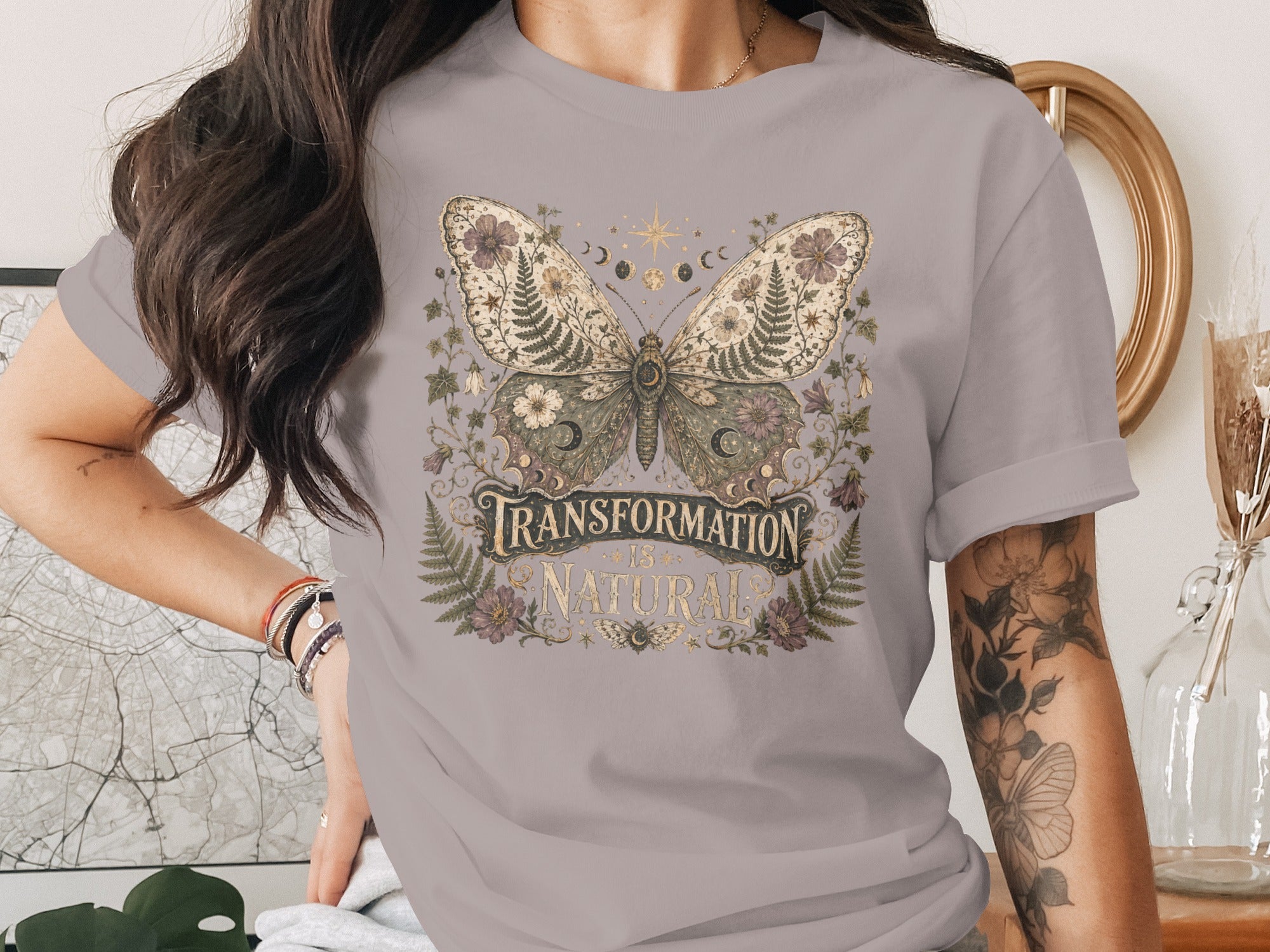 T-shirt graphic with ornate butterfly, moon phases, and text "Transformation Is Natural".