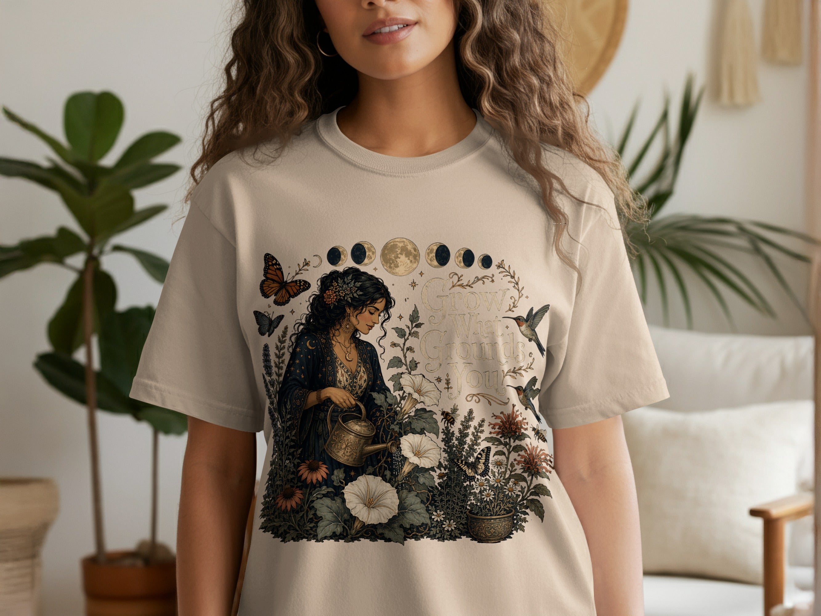 T-shirt with floral design of woman watering plants, moons, and hummingbirds. Text reads "Grow What Grounds You."