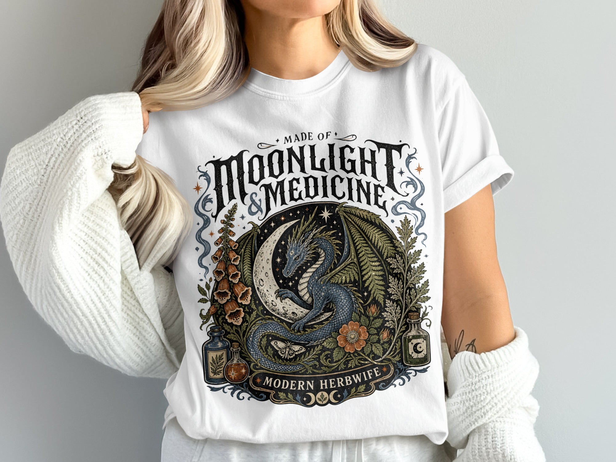 White t-shirt with a circular graphic of a blue dragon on a moon, surrounded by plants and bottles.